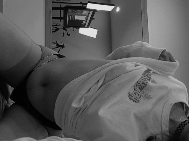 AliCipher's bare ass is visible, wearing a loose shirt, lying on a bed with a chain around her waist, legs raised, in a black-and-white photo.