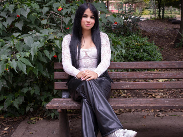DalilaTennesse with long black hair, white lace top, black leather pants, sitting on a wooden bench, wearing white sneakers, green leafy background, slight smile, hands on her knees.