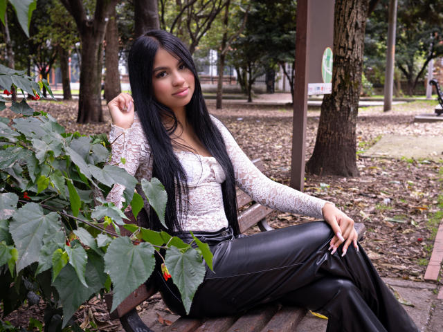 DalilaTennesse with long black hair, lace top, black leather pants, sitting on a bench, green leaves in front, right hand touching hair, left hand on knee, slight smile.