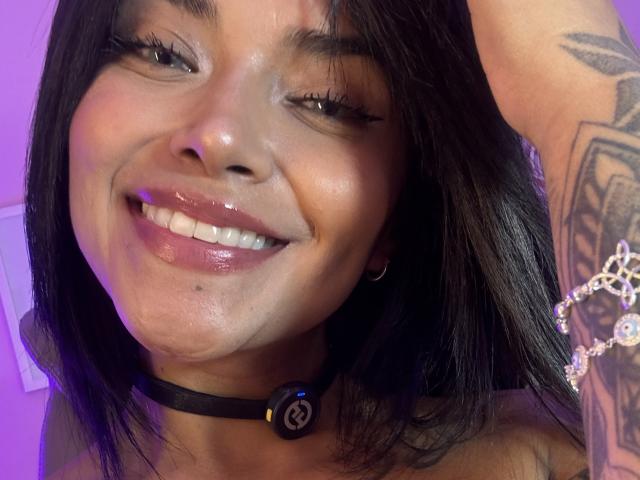MajoJones has shiny, wet-looking skin, dark hair, and is smiling with tongue out, wearing a black choker with a ring, and has a tattoo on her arm.