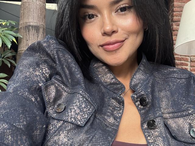 MajoJones with long black hair, wearing a snake-patterned jacket, smiling slightly, showing a cute face, and brown eyes, with a small nose and pink lips, in a selfie.
