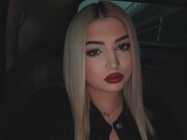 SiellaDolce tem cabelo loiro, batom vermelho escuro e sobrancelhas grossas, vestindo uma camisa preta com um botão dourado, dentro de um carro com pouca luz..