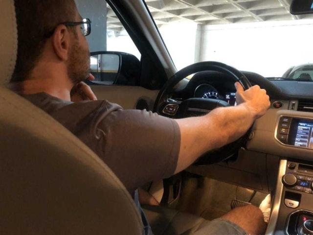 ViktorMassimo, com óculos e barba, sentado no carro, segurando o volante com a mão direita, vestindo uma camiseta cinza, assento bege, interior do carro visível, painel com display, perna esquerda cruzada..