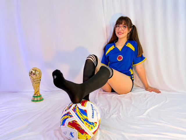 IsabelaP con coletas marrones, camiseta de fútbol azul, calcetines negros hasta las rodillas, sentada sobre un paño blanco, con las piernas levantadas, los pies sobre un balón de fútbol de colores, y el trofeo de la FIFA a la izquierda..