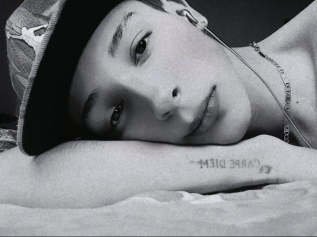 Black and white photo of MaxSteeil with a cap, lying down, head resting on arm, showing tattoo with text and symbol, wearing a chain, earbuds in.