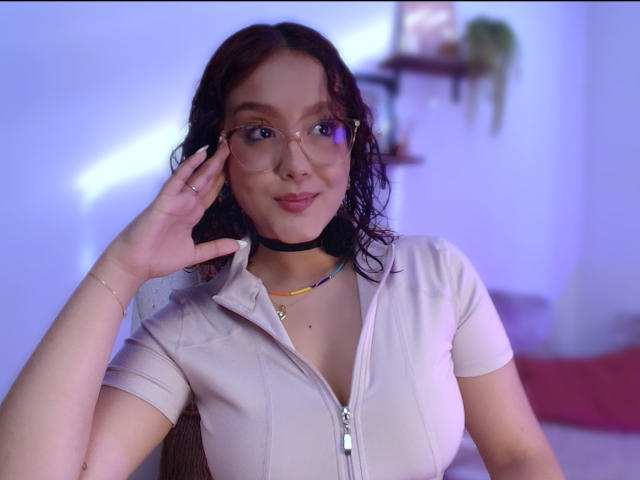 SophiaBrowm with wavy black hair, glasses, and a light zip-up shirt, touching her face, wearing a black choker and necklace, smiling subtly.