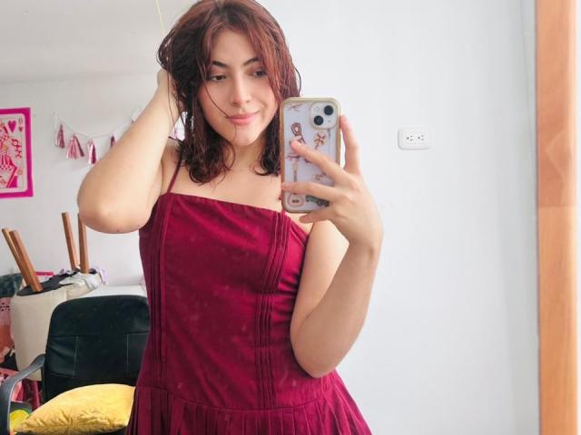 SpencerLe with wet brown hair, wearing a red strapless dress, holding a phone with a floral case, smiling, showing small tits, standing in a bright room.