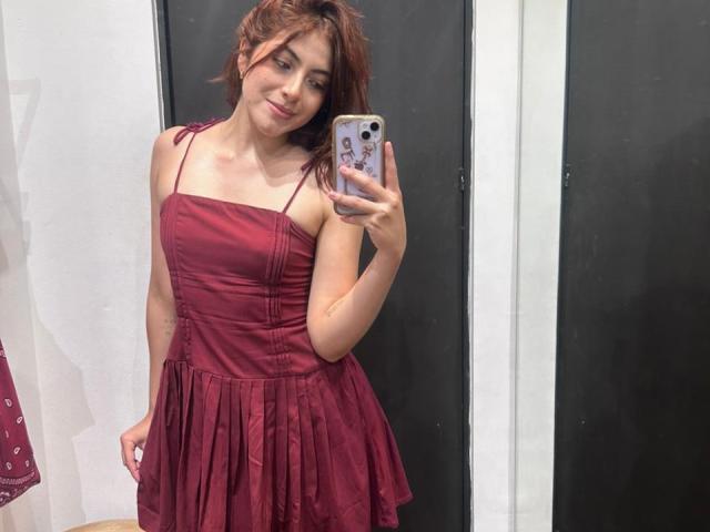 SpencerLe con un vestido rojo, sosteniendo un teléfono, sonriendo, mostrando senos pequeños, delgada, piel clara, cabello castaño, de pie frente a un espejo oscuro, en una habitación..