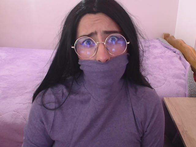 CatsitoS with long black hair, wearing round glasses with blue reflections, and a purple turtleneck that covers their nose and mouth. They look slightly confused. Purple bed in background.