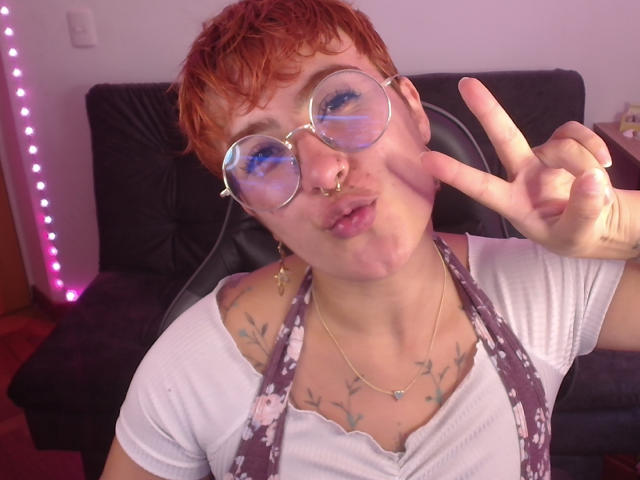 Een persoon met kort rood haar, een bril, een neusring en tatoeages, gekleed in een wit shirt en een bloemen-schort, die een vrede-teken maakt en hun borsten laat zien..