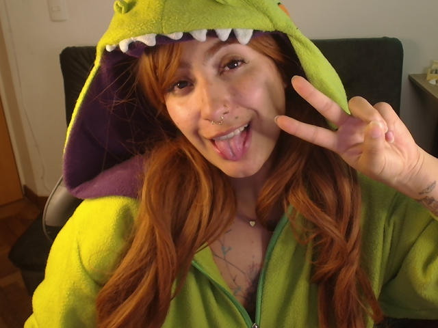 Una ragazza con i capelli rossi, felpa verde a forma di dinosauro, fodera viola, lingua fuori, piercing al naso e segno della pace, che mostra tatuaggi sul petto. Il seno è visibile attraverso la felpa aperta..