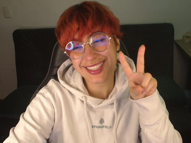 CatsitoS with red hair, glasses, nose ring, and white hoodie, making a peace sign, smiling, showing off a cute, nerdy look, with a playful, happy expression.