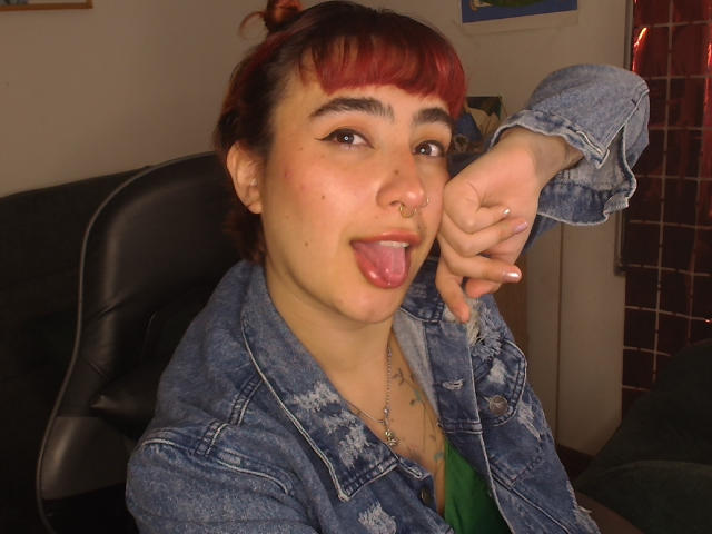 CatsitoS has red-dyed bangs, a nose ring, and is sticking out their tongue. They're wearing a blue denim jacket, green shirt, and holding up their left arm.