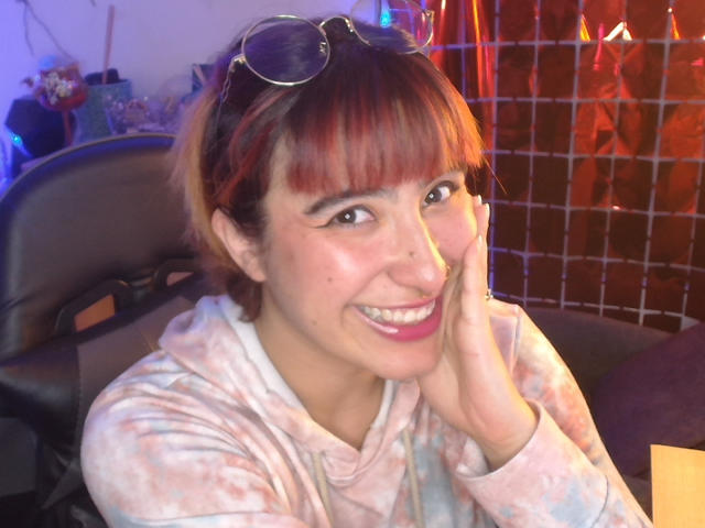 CatsitoS with red-brown hair and bangs, wearing a tie-dye hoodie, smiling with pink lipstick, eyeglasses on head, hand on cheek, sitting in a chair.