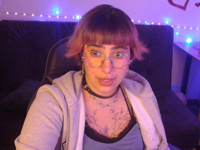 CatsitoS with short brown hair, glasses, and a choker, wearing a gray hoodie, black tank top, and showing a chest tattoo, sitting on a black sofa.