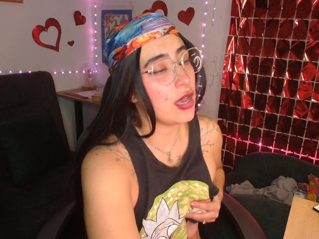 Gatos con gafas, diadema de colores, camiseta sin mangas negra, tatuajes y lápiz labial rojo, sosteniendo una rosquilla, con los ojos cerrados y la boca abierta, en una habitación tenuemente iluminada con decoraciones de corazones rojos..