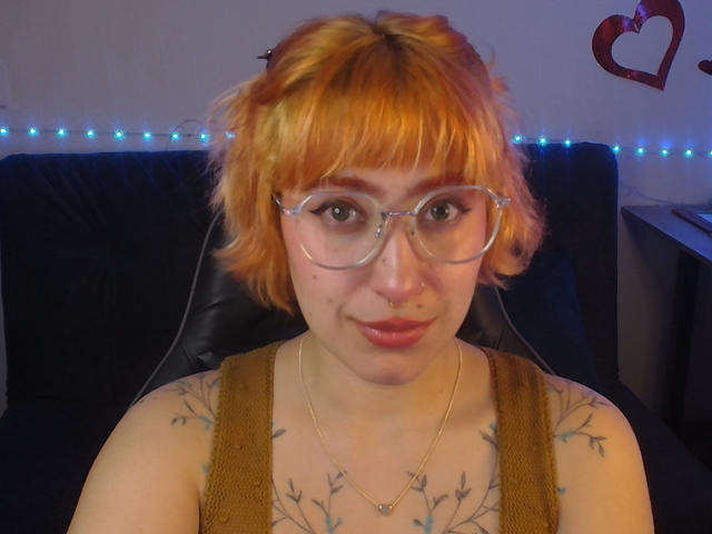 GatositoS com cabelo laranja, óculos transparentes, piercing no nariz, top marrom, tatuagens no peito, seios pequenos, sorrindo, luzes de cordão azuis, cadeira preta, decoração de coração na parede..