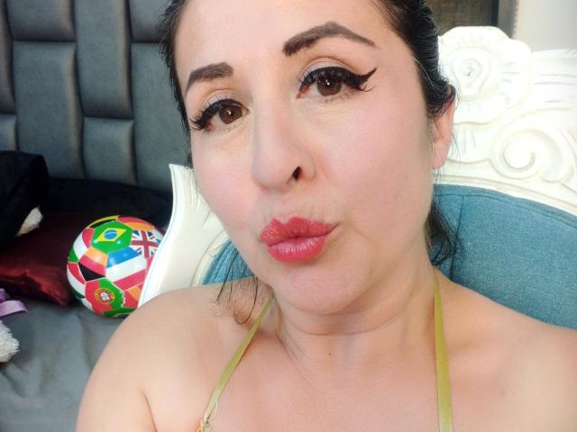 ScarletMilfX con labbra rosse carnose, eyeliner nero spesso, occhi marroni e una top verde a halter, che fa un'espressione di bacio, con una palla colorata su una sedia blu..