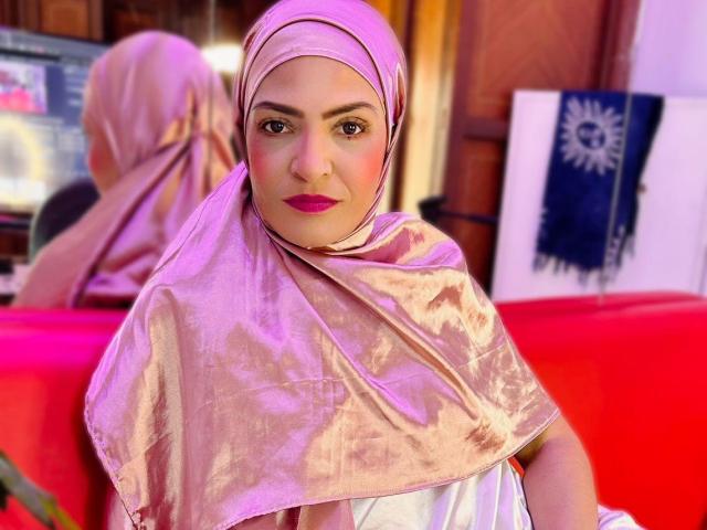 KatteRouse, con un hijab plateado rosado, un vestido blanco, lápiz labial rosa y ojos marrones, está sentada frente a una mesa roja. Su trasero está parcialmente cubierto por el hijab..