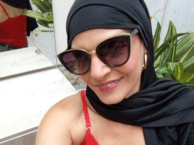 KatteRouse, con un hijab negro, un sujetador rojo, aretes de aro dorados y grandes gafas de sol negras, sonriendo y mostrando dientes metálicos. Sus pechos están cubiertos por el sujetador, y lleva lápiz labial rojo..