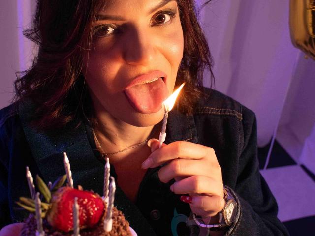 KatteRouse con la lengua afuera, lamiendo una vela de cumpleaños encendida, sosteniendo un pastel con fresas y velas, vistiendo una camisa azul oscuro y un collar dorado..