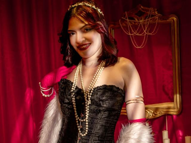 KatteRouse, con cabello rojo, un corpiño de encaje negro, guantes rosas, un collar de perlas, piel sintética blanca y una diadema de perlas, sonriendo a la cámara. Los senos son prominentes en el corpiño de encaje negro..