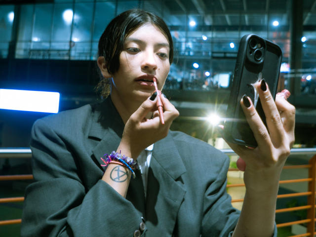 HailyEvans with short black hair, wearing a gray suit, holding a smartphone, and sucking on a red lipstick, showing a pentagram tattoo on her wrist, in a modern setting.