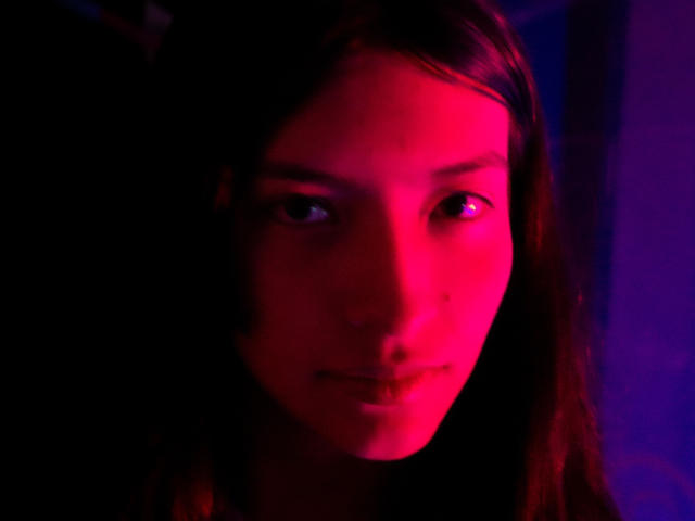 Il viso di Haily Evans è illuminato da luci rosse e viola, che mostrano un'espressione seria, lunghi capelli castani e un sorriso sottile, quasi impercettibile. I suoi occhi sono leggermente socchiusi..