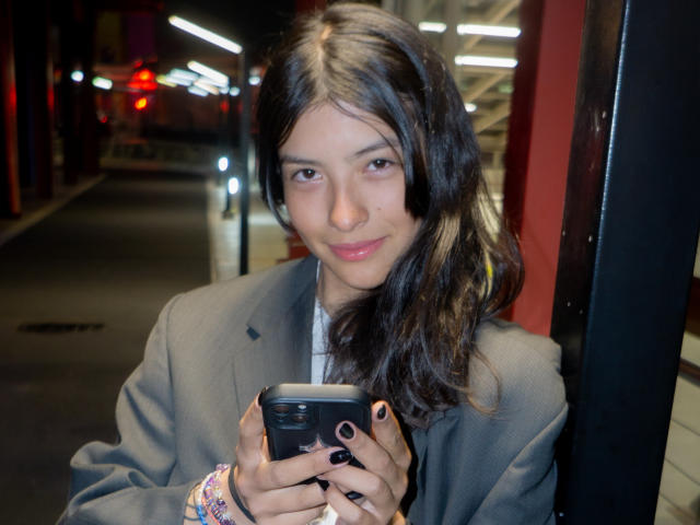 Haily Evans, con cabello oscuro y ondulado, lápiz labial rosa y esmalte de uñas negro, vestida con un blazer gris, sosteniendo una cámara, sentada en una zona urbana con poca luz, sonriendo sutilmente..