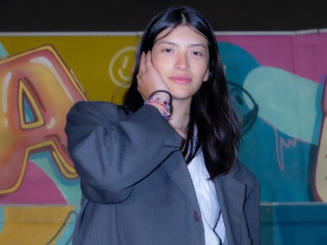 HailyEvans with long black hair, wearing a grey blazer and white shirt, hand on cheek, pink wristbands, looking slightly to the side with a calm expression.