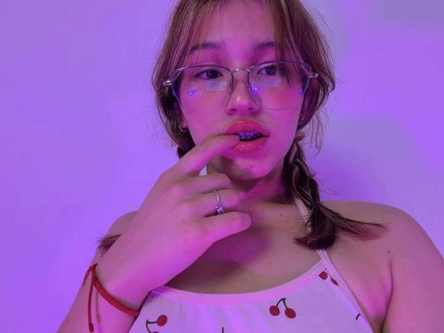Elodie Gardner, con gli occhiali, il viso illuminato di rosa e un vestito rosso a forma di ciliegia, morde un dito con un anello, indossa un braccialetto rosso e ha una treccia, mostrando il seno e le labbra rosa..