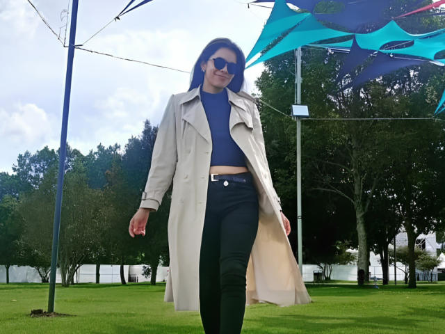Alana Blake, avec de longs cheveux bruns, un pull bleu col roulé, un pantalon noir, un trench beige et des lunettes de soleil foncées, marchant sur de l'herbe verte, sous des structures en forme de tente bleues et vertes..