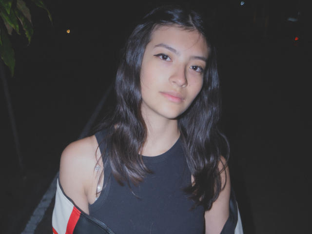 Haily Evans, con cabello negro ondulado, vestida con una camiseta negra sin mangas, una chaqueta ligeramente holgada y delineador de ojos discreto, mirando hacia un lado con una expresión neutra. Escena nocturna..