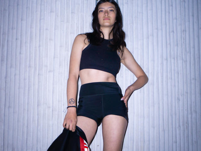 Haily Evans, com cabelo escuro, vestindo uma cropped preta e shorts pretos, segurando uma jaqueta preta com listras vermelhas, tem uma tatuagem no antebraço direito e está em pé, com uma postura confiante..