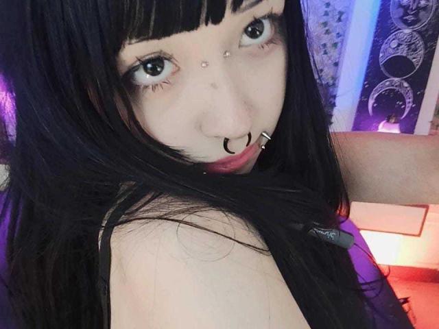 VickMoon with black hair, nose ring, septum piercing, and eye piercings, looking over shoulder, showing a red lip, in a selfie. She's wearing a black top.