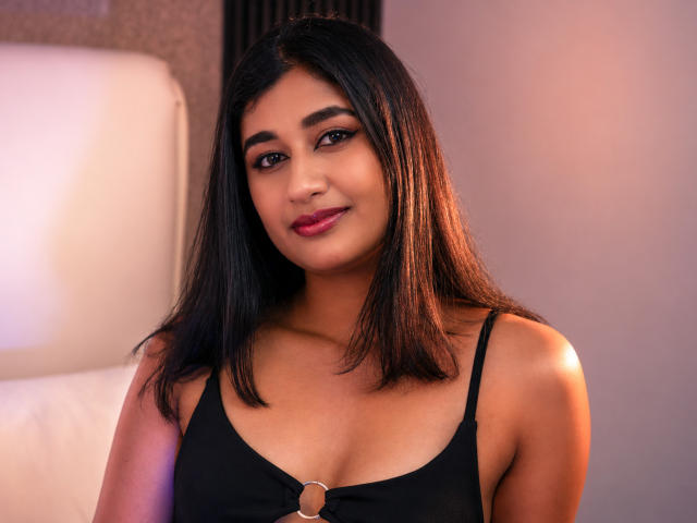 Adara Rivat, avec une peau brun clair, des cheveux noirs et lisses, et du rouge à lèvres, portant un débardeur noir avec une petite bague, souriant doucement à la caméra..