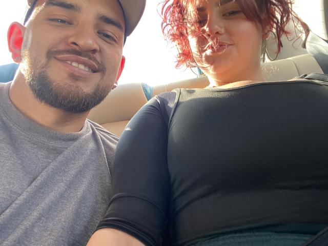 JazminYRyan with red curly hair, large tits, and black top, sitting next to JazminYRyan with brown skin, beard, and grey shirt, smiling in a car.