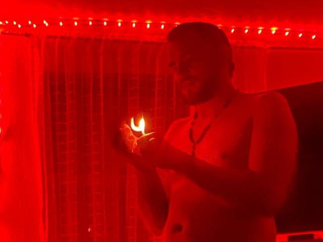 JazminYRyan, shirtless, holding a lit candle in red light, looking down, necklace, slight smile, short hair, red string lights, curtain in background.