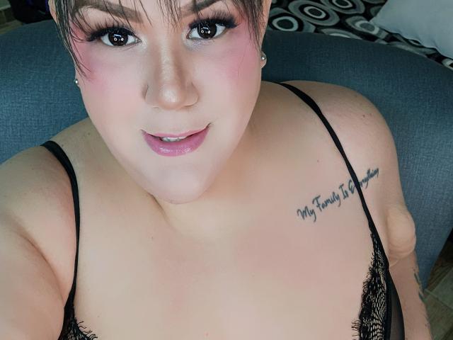 GiiannaMorrys has pink makeup, short brown hair, and a tattoo on her right shoulder saying "My Father's Little Girl". She's wearing a black lace bra, showing her tits.