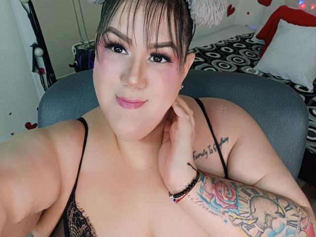 GiiannaMorrys with pink eyeshadow, black lace top, tattoos on arm and chest, smiling, hand near face, brown hair, fair skin, pink lipstick, black bra, gray chair.