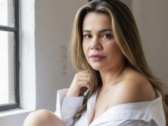 KayteHot with long brown hair, white off-shoulder shirt, and pink lipstick, looking serious, has small tits and a slight smile, sitting near a window.