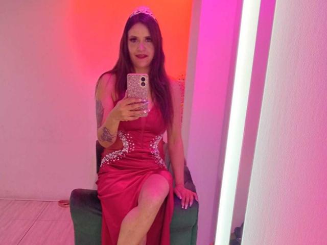 AriannaSeete in a red dress with sparkly embellishments, sitting on a green chair, holding a phone, pink and orange lights, long brown hair, tattoo on left arm, smiling.