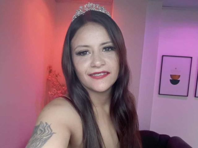 AriannaSeete with long brown hair, wearing a sparkly tiara, red lipstick, smiling, showing a tattoo on her right shoulder, in a room with pink and purple lighting.