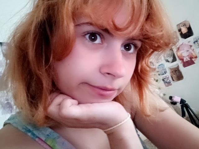 MireilleSpotwood with red, messy hair, brown eyes, and a subtle smile, resting her chin on her hand, wearing a colorful top, looking at the camera.