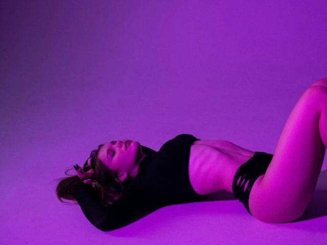 KristianSeidel is lying on their back, wearing a black top and black panties, with purple lighting highlighting their slim, flat-stomach, and visible hip bones.