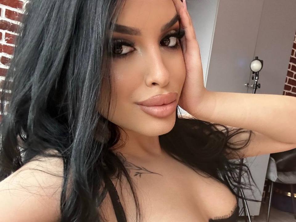 LilaVatti hat langes, schwarzes Haar, volle Lippen und ein Tattoo auf ihrer Brust. Sie berührt ihren Kopf mit ihrer rechten Hand und zeigt dabei ihre Brüste in einem schwarzen Top..