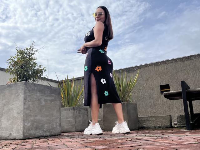 GalaMalkova, black floral dress with thigh slit, showing her ass, white sneakers, standing on brick patio, blue sky, cloudy, concrete walls, planters, smiling, looking back.