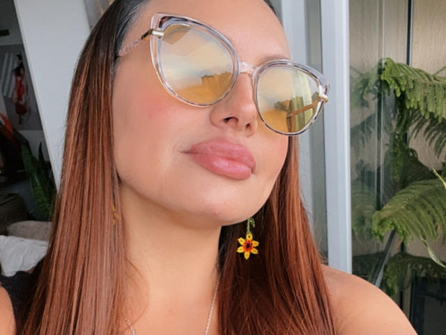 Gala Malkova mit glänzendem, braunem Haar, die klare Sonnenbrillen trägt, die das Licht reflektieren, hat rosige, glänzende Lippen, ein kleines, gelbes Blumen-Ohrring und einen leichten Schmollmund, und sieht dabei niedlich aus..