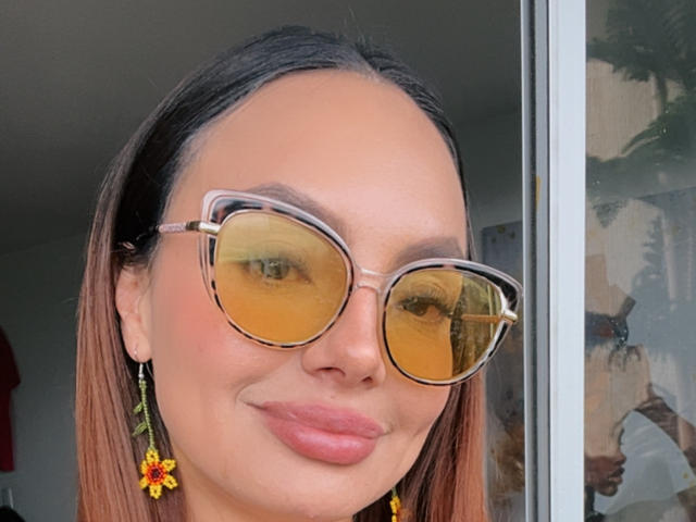 GalaMalkova, con i capelli castani, indossa occhiali a forma di gatto di colore giallo, rossetto rosa e orecchini colorati a forma di fiore, e sorride dolcemente. La sua pelle è abbronzata e appare carina e alla moda..