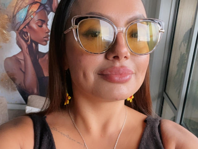 GalaMalkova, con la pelle abbronzata, i capelli castani, indossa occhiali con montatura gialla, orecchini gialli e una maglietta nera, mostrando un piccolo tatuaggio sulla clavicola destra e un sorriso appena accennato..